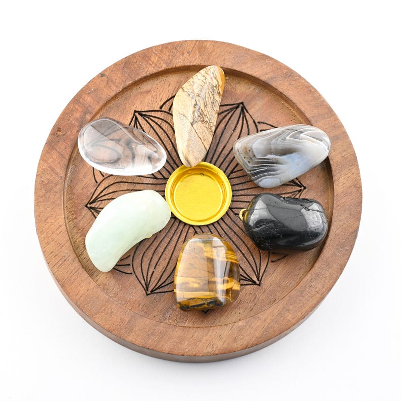 May include: A wooden tray with a floral design holds seven polished gemstones of various colors and patterns. The gemstones include clear, brown, green, black, and tiger's eye stones. A gold-colored circular object is in the center.