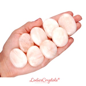 Op de afbeelding: Een hand die acht gladde, ovale, lichtroze getrommelde stenen vasthoudt. De stenen zijn gepolijst en hebben een lichte glans. De stenen zijn in een waaier vorm gerangschikt, met de grootste steen bovenaan en de kleinste steen onderaan. De stenen zijn allemaal ongeveer even groot en even gevormd. De achtergrond is wit.