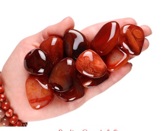 May include: A hand holding a collection of carnelian tumbled stones. The stones are a deep red color with some orange and brown hues. The stones are smooth and polished.