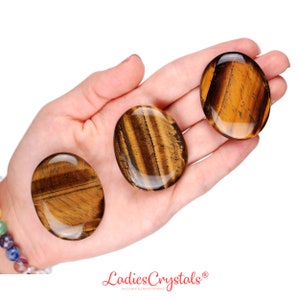 May include: Three polished tiger's eye stones, oval-shaped, are displayed in a hand. The stones have a rich, golden-brown color with distinctive bands and patterns. The stones are smooth and glossy, reflecting light.