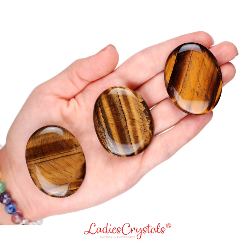May include: Three polished tiger's eye stones, oval-shaped, are displayed in a hand. The stones have a rich, golden-brown color with distinctive bands and patterns. The stones are smooth and glossy, reflecting light.