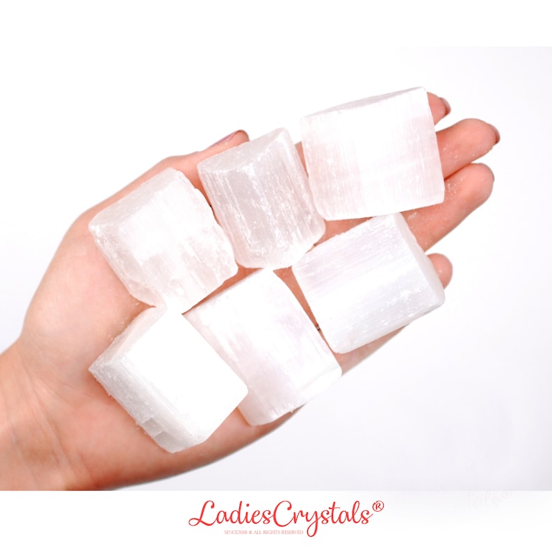 May include: Six white selenite crystal cubes in a hand. The crystals are smooth and have a striated texture.