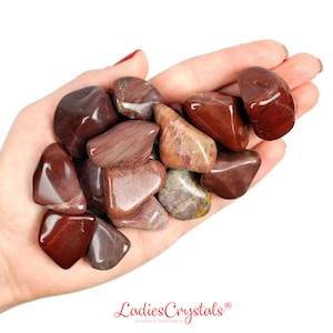 Puede incluir: Un puñado de piedras de jaspe rojo pulidas. Las piedras varían en forma y tamaño, con una mezcla de superficies sólidas y estampadas. Los colores van desde el burdeos intenso hasta los tonos marrón rojizo más claros. La imagen incluye el texto "Ladies Crystals".