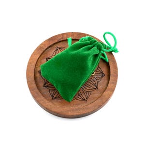May include: A round, wooden tray with a carved floral design holds a small, emerald green velvet drawstring bag. The bag is closed with a matching green cord. The tray is a warm brown color.