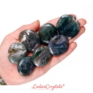 May include: A collection of polished moss agate stones in varying shades of green, white, and gray. The stones are oval-shaped and held in a hand, showcasing their natural patterns and textures. The image includes the text "Ladies Crystals".