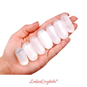 May include: Seven smooth, white selenite tumbled stones in the shape of an oval. The stones are arranged in a line in a hand.