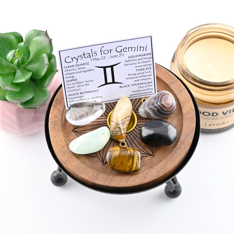 May include: A wooden tray with a variety of gemstones, including clear quartz, jasper, and tiger eye. A card reads "Crystals for Gemini" with descriptions of each stone. A small succulent plant and a peach-scented candle are also in the image.