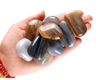 Pode incluir: Uma coleção de pedras de ágata polidas em tons de castanho, cinza e azul. As pedras são lisas e ovais, com padrões de bandas visíveis. A imagem inclui o texto "Ladies Crystals®" na parte inferior.