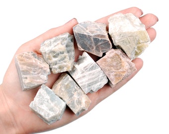 May include: A collection of eight rough, natural, gray and pink feldspar crystals. The crystals are various sizes and shapes, and have a rough, natural texture.