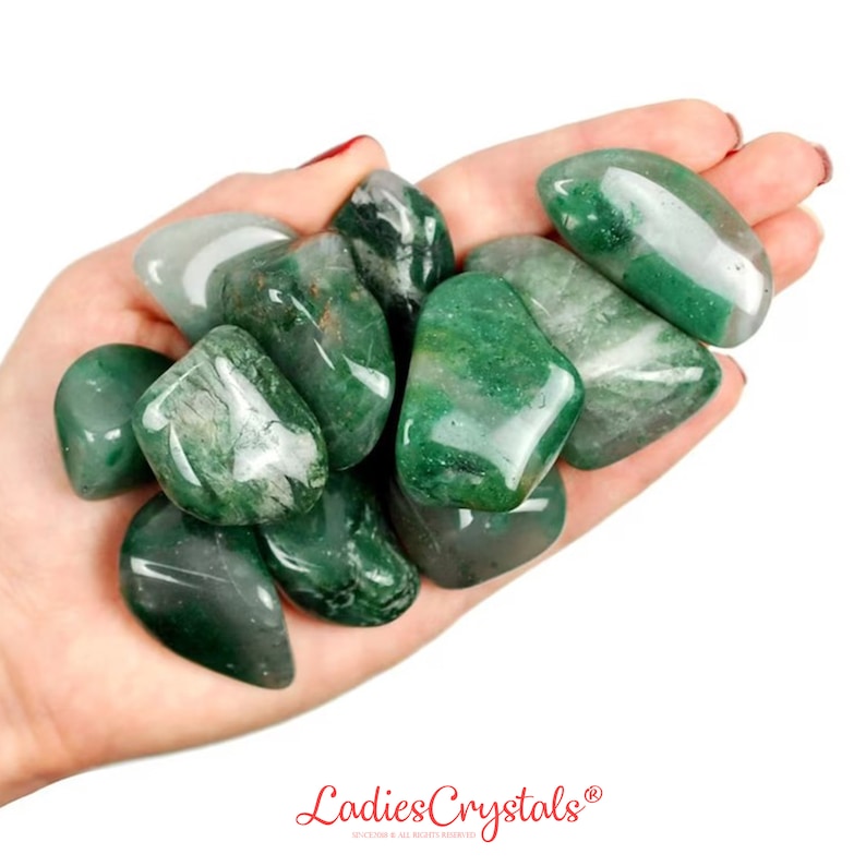 May include: A handful of polished green aventurine stones. The stones vary in shades of green, from light to deep forest green, with some displaying white veining. The stones are smooth and rounded, with a glossy finish. The logo "Ladies Crystals" is visible at the bottom.