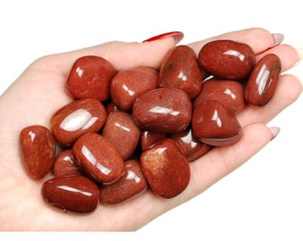 May include: A collection of polished, reddish-brown gemstones, oval-shaped, and smooth, held in a hand. The stones have a glossy finish, reflecting light. The image includes the text "Ladies Crystals".