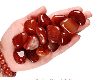 May include: A hand holding a pile of red carnelian tumbled stones. The stones are smooth and polished, with a deep red color.