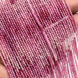 Peut inclure: Plusieurs brins de petites perles roses rondes. Les perles varient en teinte, du rose clair au rose foncé, créant un effet de dégradé. Les perles sont enfilées ensemble, formant de nombreux brins. Le fond est blanc.