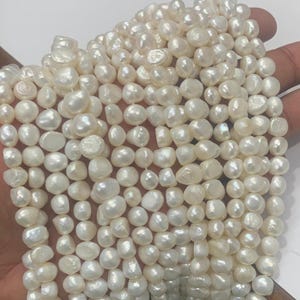 Può includere: Fili di perle d'acqua dolce bianco crema. Le perle hanno forma e dimensioni irregolari, con una superficie lucida. I fili sono tenuti in una mano, mostrando la bellezza naturale delle perle. Ideale per la creazione di gioielli.