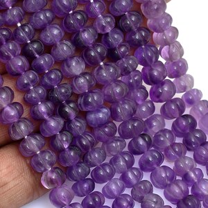 Peut inclure: Un collier de perles d'améthyste violettes rondes présentant une texture sculptée et nervurée. Les perles sont enfilées en plusieurs lignes parallèles, révélant des nuances variées de violet translucide et de pourpre profond dans la pierre.