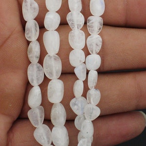 Può includere: Primo piano di fili di perline di pietra di luna bianche. Le perline di forma ovale sono infilate insieme, formando una collana o un braccialetto. Le perline hanno una qualità traslucida e una lucentezza sottile.