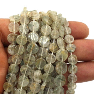 Peut inclure: Plusieurs rangées de perles rondes translucides, de couleur vert pâle et beige. Les perles sont enfilées, idéales pour un collier ou un bracelet. Les perles sont tenues dans une main sur fond blanc.