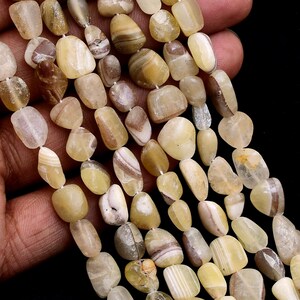 Può includere: Più fili di perline levigate di forma irregolare in tonalità crema, beige, sabbia e marrone chiaro. Le perline presentano superfici lisce e lucide con sottili venature naturali e aree traslucide, infilate su un sottile cordino bianco.