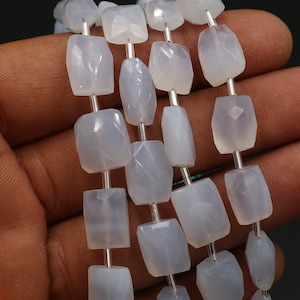 Puede incluir: Primer plano de cuentas de piedras preciosas facetadas de color azul claro. Las cuentas son rectangulares y están ensartadas en un alambre plateado. Las piedras preciosas son translúcidas. Fondo oscuro.