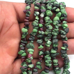 Può includere: Un filo di perline di pietre preziose verdi e viola. Le perline sono piccole e di forma irregolare, simili a schegge o ciottoli. Il filo è composto da più file di perline, creando un effetto a strati.