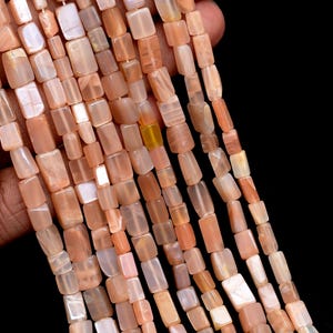 May include: Multiple strands of rectangular, polished peach-coloured moonstone beads. Each bead features a smooth, semi-translucent surface with subtle variations in tone, ranging from pale cream to deep orange, strung on thin white thread.