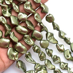 Op de afbeelding: Olijfgroene onregelmatig gevormde kralen geregen op witte draad. De kralen hebben een glanzende afwerking en worden in een hand gehouden tegen een witte achtergrond. De kralen zijn een mix van organische vormen en maten, geschikt voor het maken van sieraden.