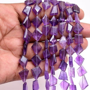 Può includere: Una collezione di perline in ametista sfaccettate disposte in più fili verticali. Ogni perlina presenta una distinta forma geometrica a rombo o aquilone con una tonalità viola traslucida, mostrando variazioni naturali nel colore e nella chiarezza.