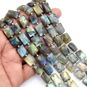 Può includere: Fili di perline rettangolari di labradorite nelle tonalità del grigio, blu e giallo. Le perline sono infilate insieme, mostrando le loro superfici sfaccettate e il gioco di colori iridescenti. Le perline sono tenute in una mano.