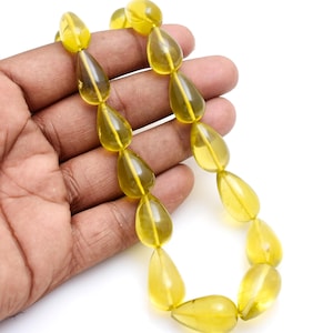 May include: A necklace of translucent, teardrop-shaped beads in shades of yellow and amber. The beads are strung together, creating a flowing, organic design. The necklace is held in a hand against a white background, showcasing the color and shape of the beads.