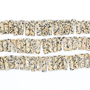 Könnte beinhalten: Reihen rechteckiger Dalmatiner-Jaspis-Perlen. Die Perlen sind hellbeige mit schwarzen Flecken. Die Perlen sind aufgereiht und bereit für die Schmuckherstellung. Die Perlen sind etwa 2,5 cm lang.