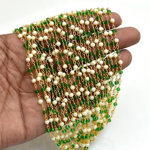 Op de afbeelding: Meerdere strengen kralen, afwisselend wit en groen, geregen op goudkleurige kettingen. De strengen worden in een hand gehouden tegen een witte achtergrond. De kralen zijn klein en rond. Decoratief geheel.