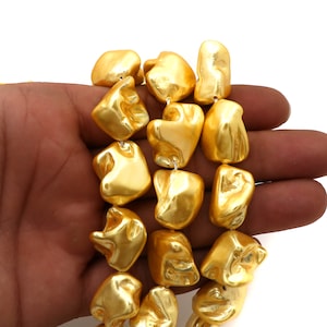 Op de afbeelding: Een streng grote, onregelmatige gouden kralen. De kralen hebben een glanzende, metallic afwerking en zijn aan elkaar geregen. De kralen worden in een hand gehouden, wat hun grootte en vorm laat zien. De kralen lijken van een solide materiaal te zijn gemaakt.
