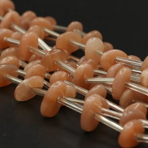 Könnte beinhalten: Eine Strähne aus pfirsichfarbenen Sunstone-Perlen, die auf einem klaren Glasdraht aufgefädelt sind. Die Perlen sind oval geformt und haben einen leicht schimmernden Schimmer.