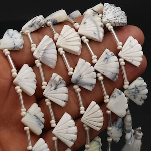 Puede incluir: Primer plano de un collar con cuentas blancas en forma de abanico con vetas grises. Las cuentas están ensartadas con pequeñas cuentas blancas y varillas plateadas. El collar se muestra sobre un fondo oscuro.