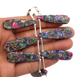 May include: A collection of elongated, teardrop-shaped gemstone beads. The beads are a mosaic of colors, including red, blue, green, and gold, with silver metal accents. The beads are approximately 1.5 inches long.