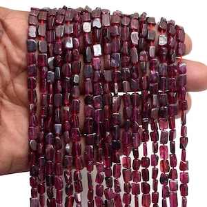 Peut inclure: Plusieurs brins de perles de grenat rouge foncé. Les perles sont rectangulaires et enfilées sur un fil rouge. Les perles ont une surface polie, légèrement irrégulière. L'image montre plusieurs brins de perles, tenus dans une main.