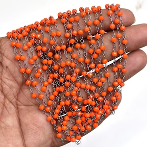 May include: A long jewelry chain featuring small, faceted, bright orange beads spaced evenly along a dark metallic wire. The chain is draped in multiple parallel rows, showcasing the vibrant color of the beads against the dark, thin metal links.