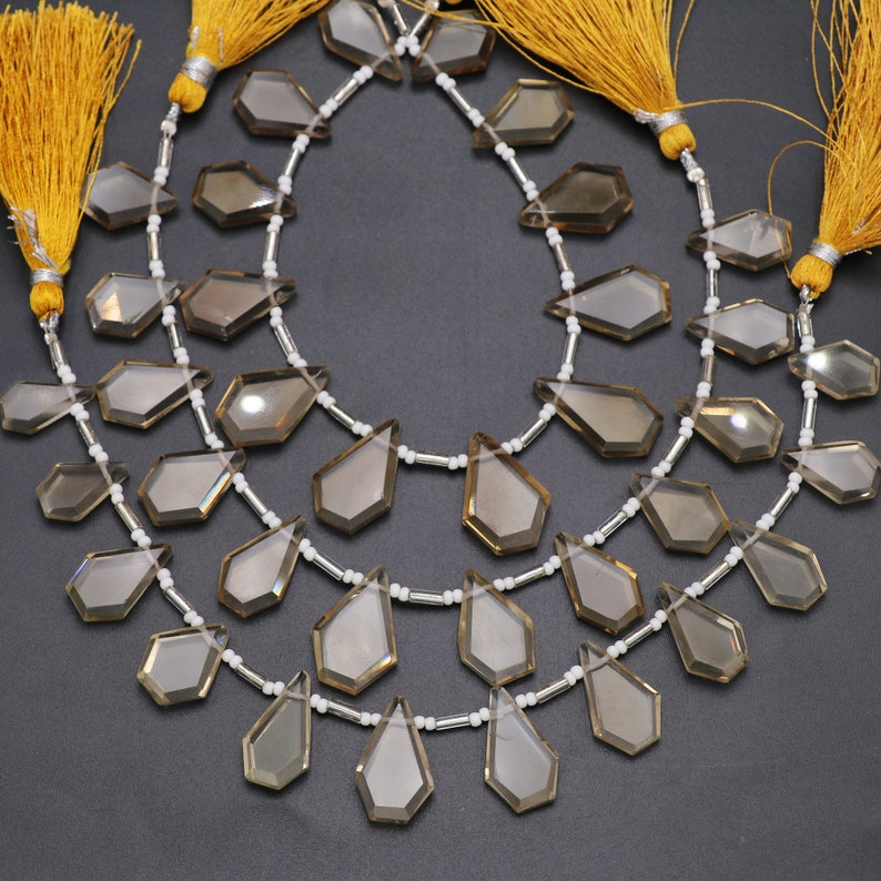 Puede incluir: Una hebra de cuentas de piedra preciosa de cuarzo ahumado con formas de l&aacute;grima facetadas. Las cuentas est&aacute;n enhebradas en un alambre plateado con cuentas espaciadoras blancas. La hebra est&aacute; terminada con dos borlas amarillas.
