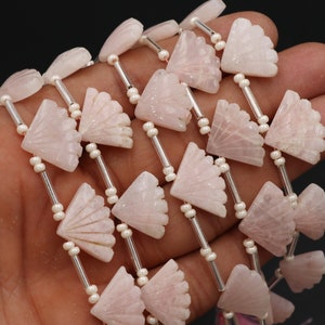 Peut inclure: Gros plan sur un rang de perles de quartz rose. Les perles sont sculptées en forme d'éventail et enfilées avec de petites perles blanches et des tubes métalliques argentés. Le fond est sombre.