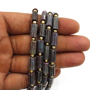 以下が含まれることがあります： 宝石ビーズの複数のストランドのクローズアップ。ビーズは濃い赤紫の色で、金色のスペーサーが付いています。ビーズは円筒形で、ファセット加工された表面を持っています。背景は白です。