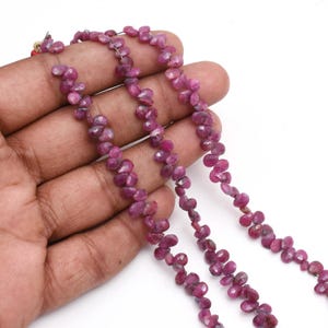 Peut inclure: Plusieurs brins de perles de rubis en forme de goutte, facettées, de différentes nuances de rose et de rouge. Les perles sont enfilées, créant une apparence délicate et élégante. Les pierres précieuses sont présentées sur fond blanc.