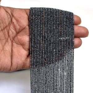 Peut inclure: Plusieurs brins de petites perles gris foncé sont tenus dans une main sur fond blanc. Les perles sont enfilées, créant un effet texturé et scintillant. Les perles semblent être de taille et de forme uniformes, possiblement des pierres précieuses.