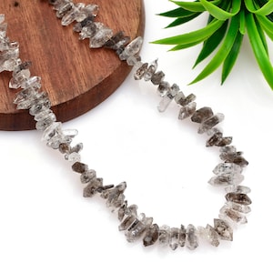 Puede incluir: Un collar formado por una hilera de cristales de cuarzo ahumado en bruto y sin tallar. Las piedras individuales son translúcidas con tonos variados de gris, marrón y cuarzo transparente, presentando bordes irregulares y puntas naturales.