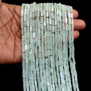 Può includere: Fili di perline rettangolari verde chiaro. Le perline sono infilate con un filo sottile verde chiaro. Le perline sono tenute nella mano di una persona su uno sfondo nero. Le perline sono di un colore verde pallido e traslucido.