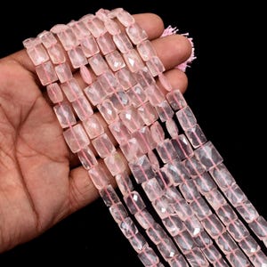 Könnte beinhalten: Stränge aus facettierten, rechteckigen Rosenquarzperlen. Die Perlen haben eine zarte rosa Farbe und sind auf rosa Faden aufgefädelt. Die Perlen werden in einer Hand vor schwarzem Hintergrund gehalten. Ideal für die Schmuckherstellung.