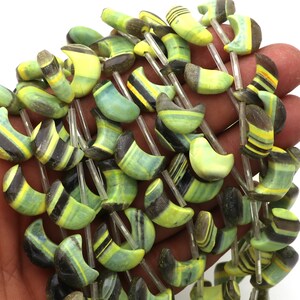 Può includere: Una collana di perline a strisce verdi, gialle e nere. Le perline sono a forma di mezzaluna e infilate su cordoncini sottili e trasparenti. Le perline sono tenute in mano su sfondo bianco.