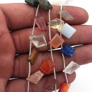 Op de afbeelding: Een streng zilveren ketting met verschillende edelsteen kralen, waaronder heldere, roze, oranje, bruine, blauwe en groene. De kralen zijn gefacetteerd en hebben verschillende vormen, waaronder vierkant, rechthoekig en driehoekig.