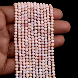Può includere: Un filo di piccole perline rotonde rosa pallido con sottili variazioni di tono e consistenza. Le perline sono infilate in una linea verticale, mostrando una finitura liscia e lucida e una forma sferica costante lungo tutta la lunghezza.