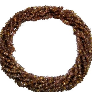Könnte beinhalten: Stränge aus kleinen, unregelmäßig geformten Edelsteinen in Braun-, Bernstein- und Olivgrüntönen. Die Edelsteine sind zu einer kreisförmigen Form auf weißem Hintergrund aufgereiht. Die Edelsteine scheinen natürlich zu sein und haben eine leicht raue Textur.