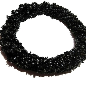 Op de afbeelding: Een zwarte kralenketting bestaande uit kleine, onregelmatig gevormde stenen. De ketting is cirkelvormig, met de kralen dicht op elkaar geregen. De kralen hebben een glanzende afwerking, die licht reflecteert tegen een witte achtergrond. De sieraden zijn geschikt voor dagelijks gebruik.